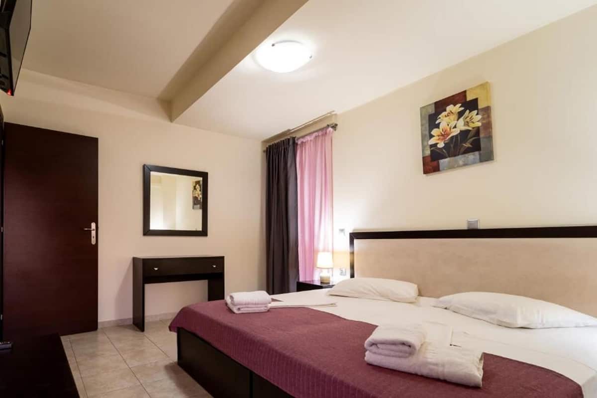 A hotel room with a bed, a bedside table, a mirror, a painting, and a window with curtains.