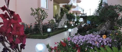 Un edificio residencial con varios balcones, un jardín bien cuidado con diversas flores y un camino pavimentado.