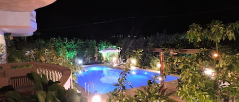 Vista nocturna de una zona de piscina con un pabellón, rodeada de vegetación y una terraza bien iluminada.