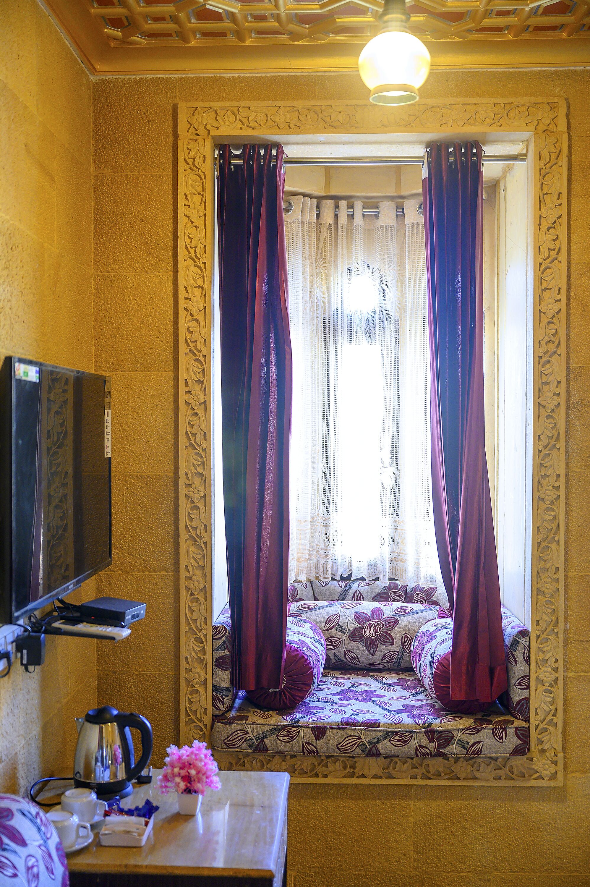 A room with a sofa bed, a television, a table with a kettle and flowers, and a window with curtains.