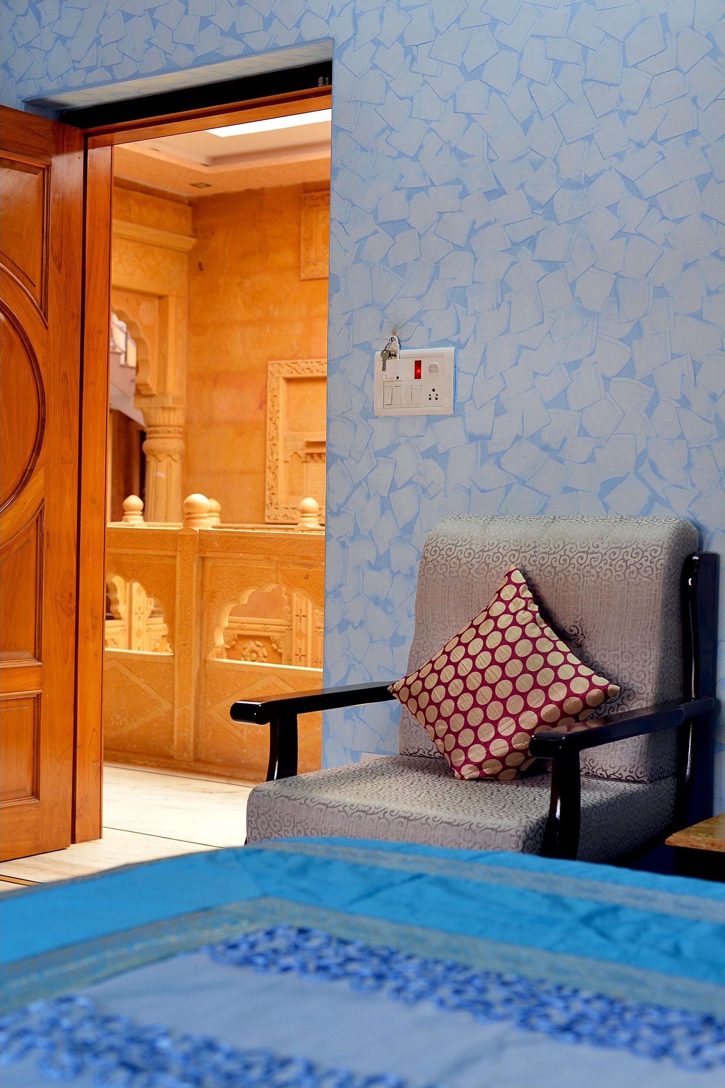 Una habitación de hotel con un papel tapiz estampado, una puerta de madera, un sillón gris con un cojín estampado y una cama con colcha azul y blanca.