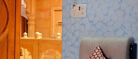 Una habitación de hotel con un papel tapiz estampado, una puerta de madera, un sillón gris con un cojín estampado y una cama con colcha azul y blanca.