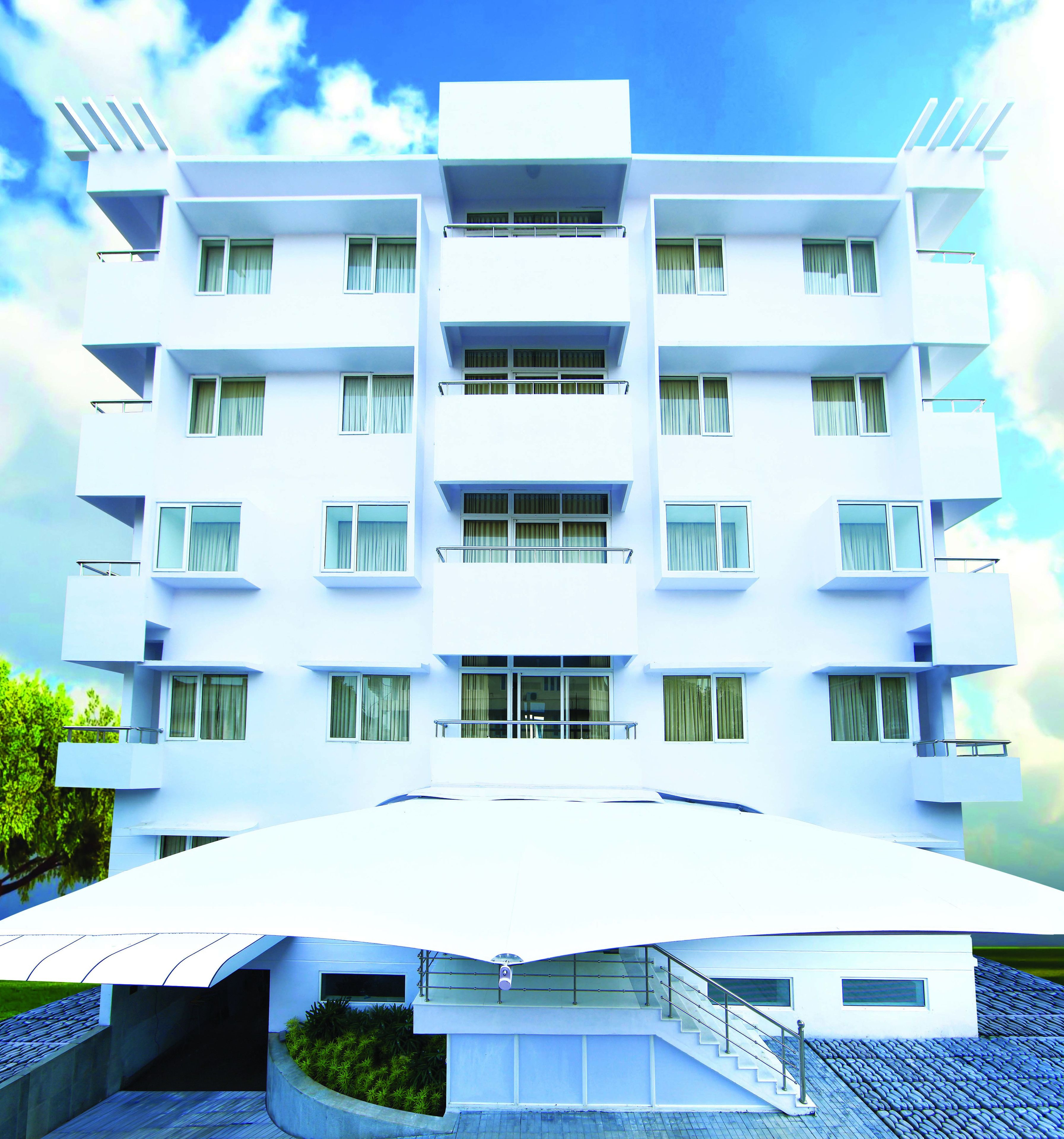 A modern white building with multiple balconies and windows, featuring a flat roof and a covered entrance area.