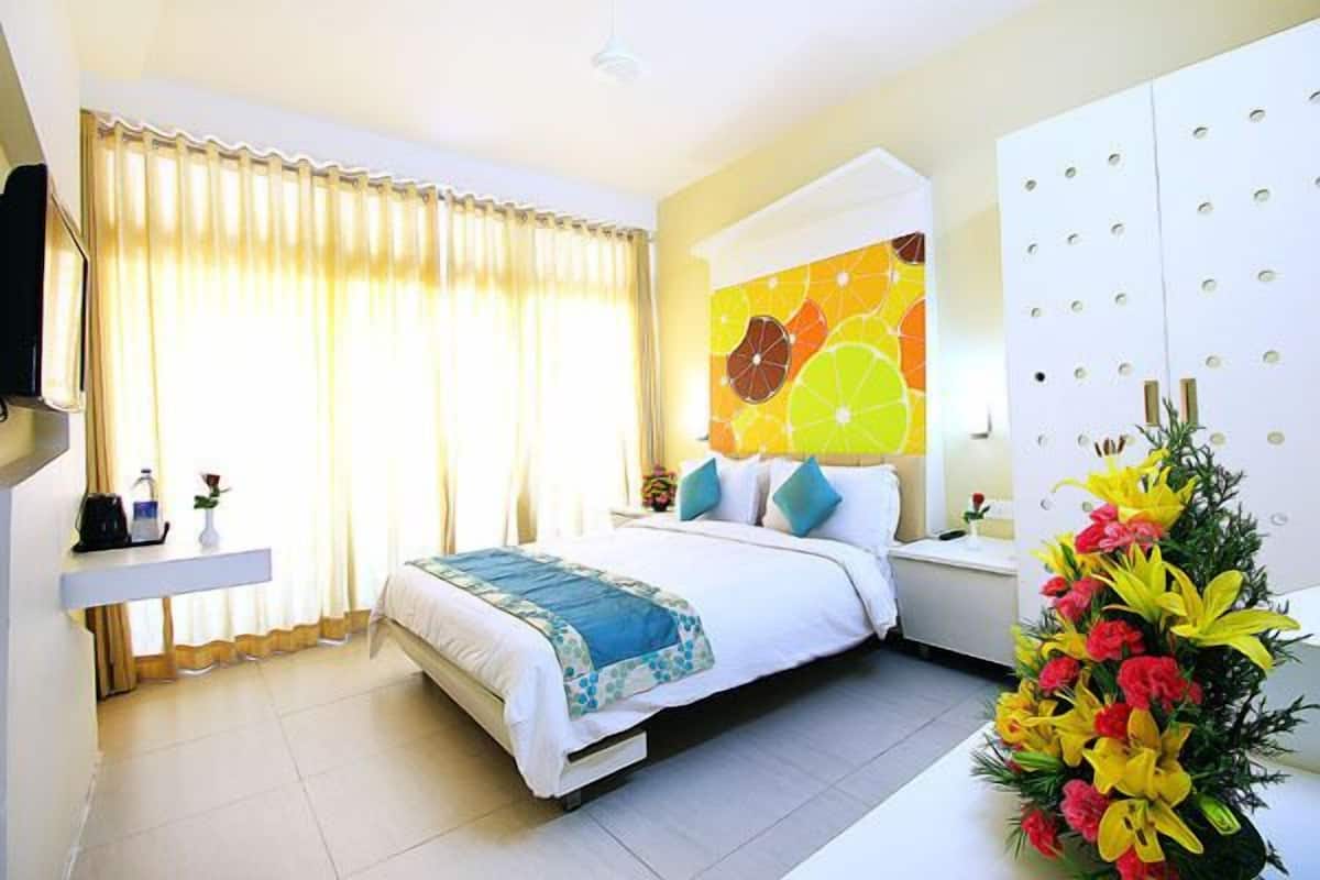 Habitación de hotel con una cama, una mesita de noche, un televisor, una ventana con cortinas y un papel tapiz con motivos de frutas.
