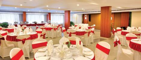 Un salón de banquetes con mesas redondas dispuestas para el servicio, cada una con mantel blanco y fajín rojo, y sillas blancas adornadas con fajines rojos.