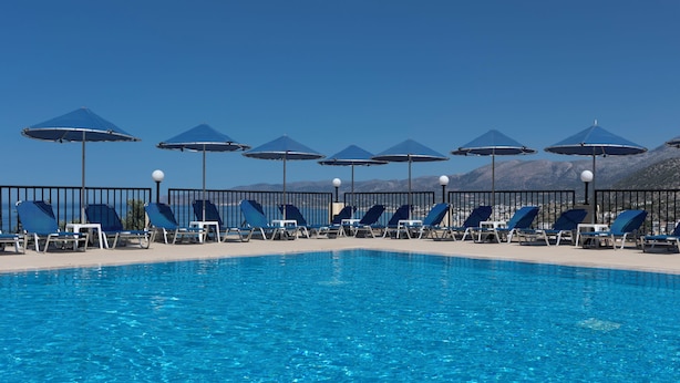 Piscina com água azul, cercada por espreguiçadeiras e guarda-sóis, com montanhas ao fundo.
