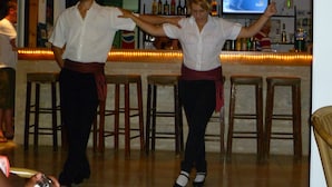 Uma área de bar com banquetas de madeira, um balcão com garrafas e uma tela de televisão.