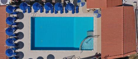 An aerial view of a swimming pool surrounded by blue umbrellas and lounge chairs, with a red-roofed building nearby.