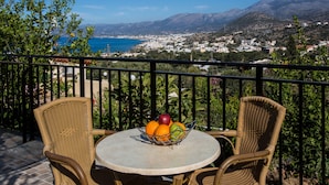 Varanda com cadeiras de vime e uma mesa, uma cesta de frutas e uma vista panorâmica de uma cidade costeira e montanhas.