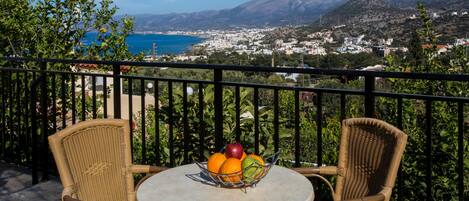 Un balcón con sillas de mimbre y una mesa, un frutero y una vista panorámica de un pueblo costero y montañas.