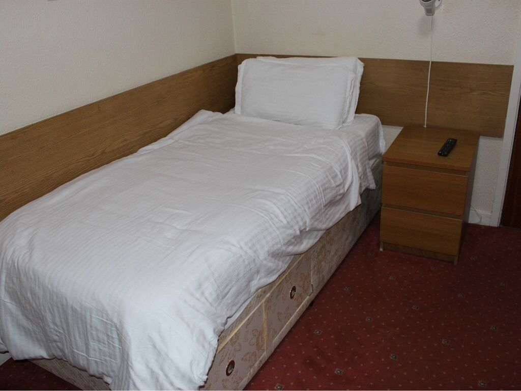 A single bed with a wooden headboard, a white mattress, and a wooden nightstand with a remote control.