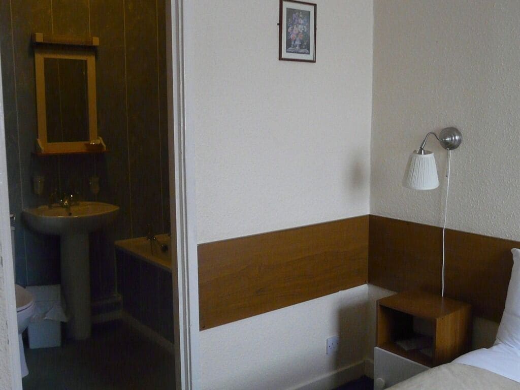 Une chambre d’hôtel avec une tête de lit en bois, une table de chevet, une lampe, un petit tabouret en bois et une salle de bain équipée d’un lavabo et de toilettes.