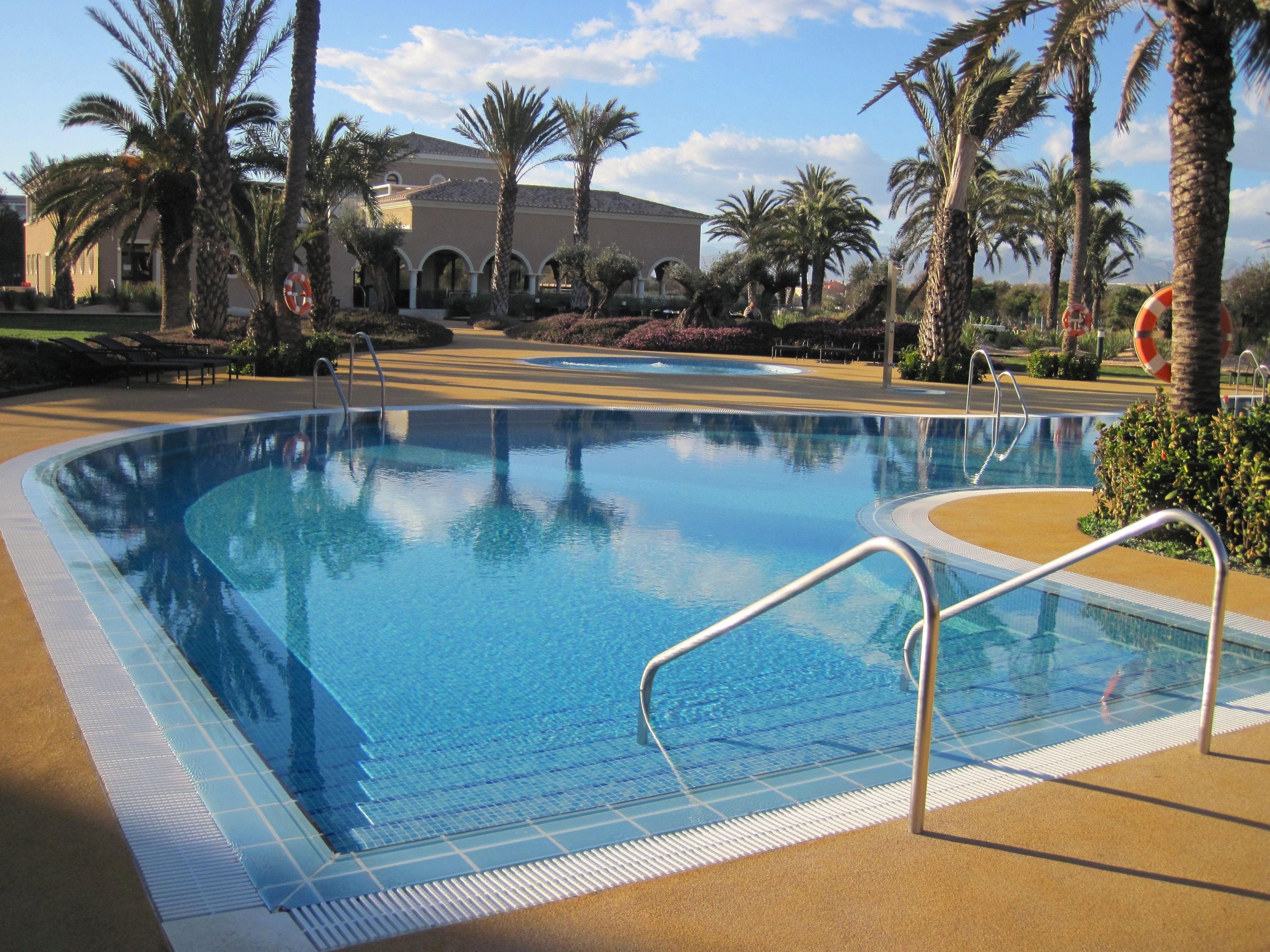 Une piscine dont le bord est revêtu de carreaux, entourée de palmiers et, au fond, un bâtiment orné d’arches.