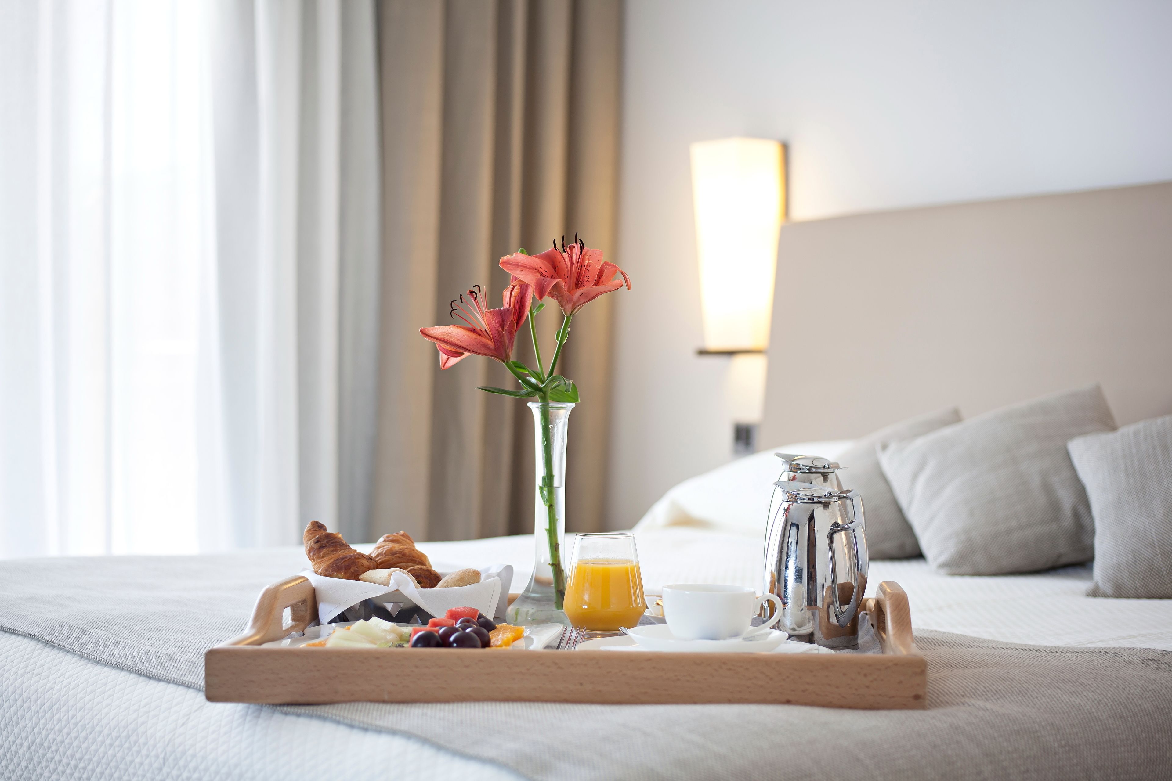 A breakfast tray with croissants, fruits, and orange juice on a bed with a vase of flowers and a lamp in the background.
