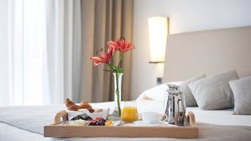 A breakfast tray with croissants, fruits, and orange juice on a bed with a vase of flowers and a lamp in the background.