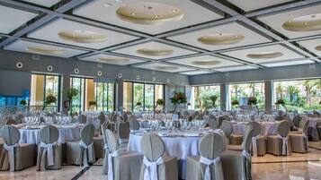A large banquet hall with multiple round tables set for dining, featuring white tablecloths and grey chairs.