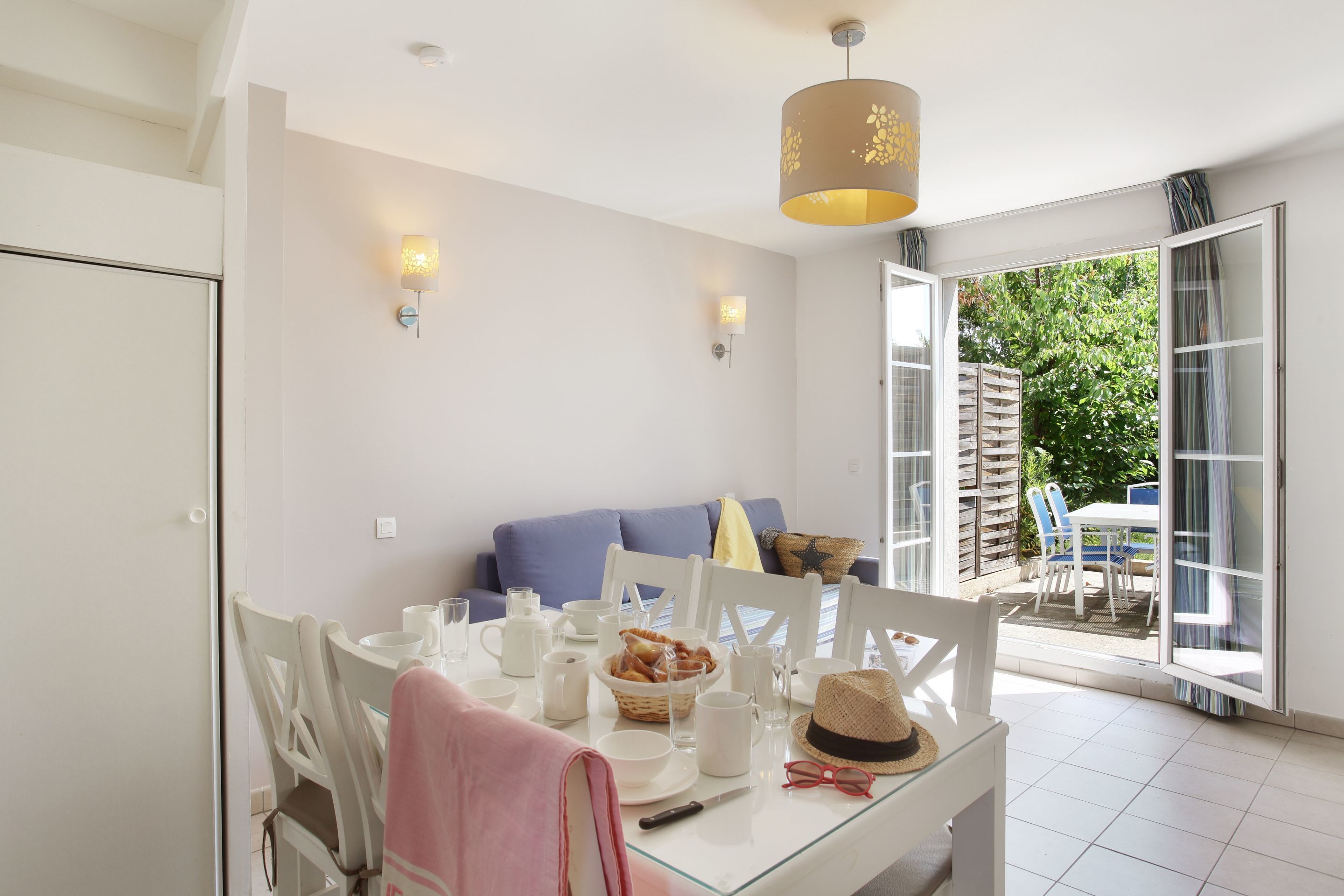 A dining area with a glass table, white chairs, and a blue sofa. There is a basket of bread on the table, and a hat and glasses on the right side.