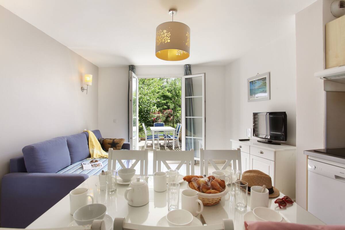 Una cocina moderna con electrodomésticos blancos, una mesa de comedor dispuesta para el desayuno y vistas del jardín a través de una puerta abierta.