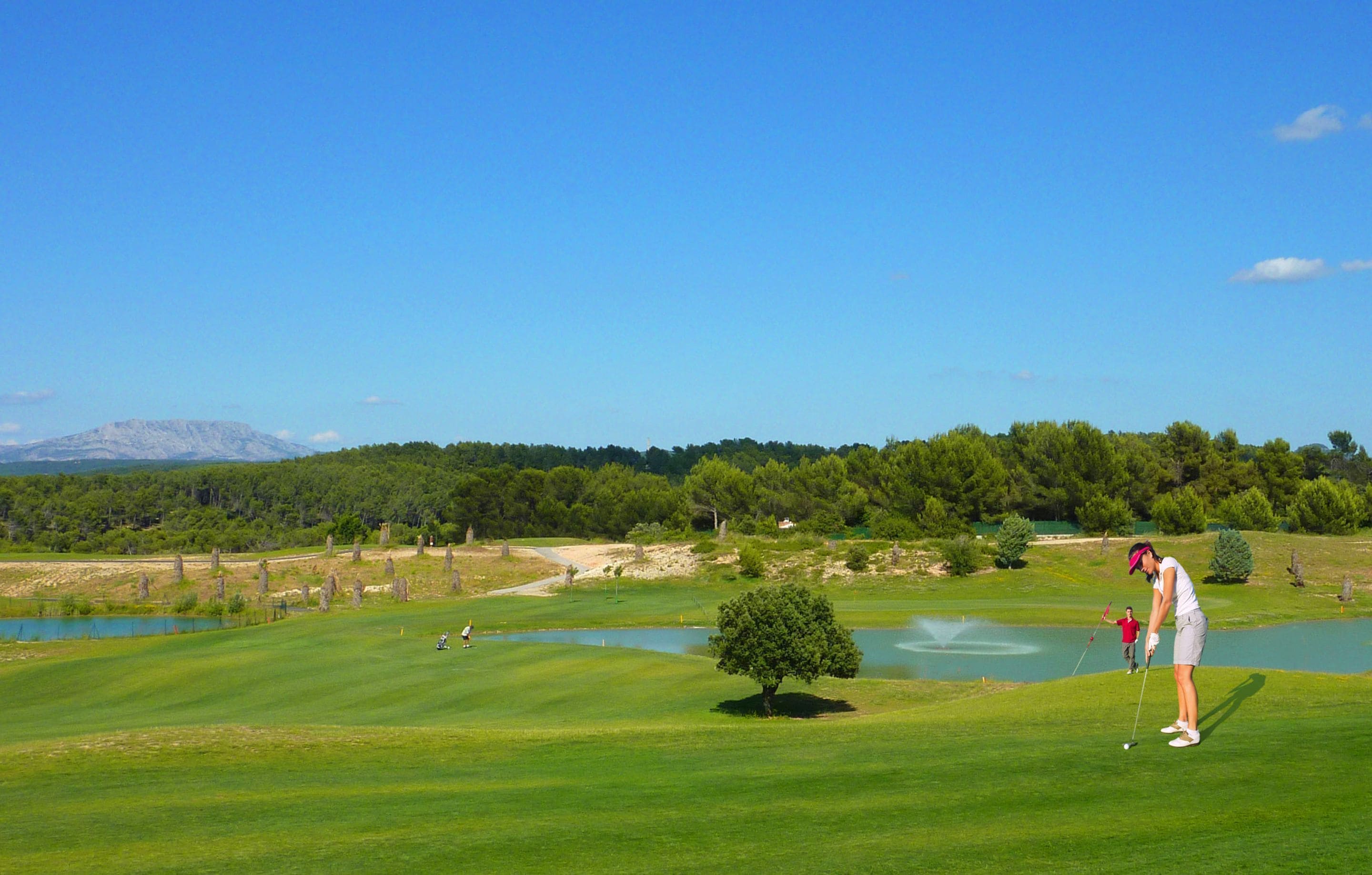 Un campo de golf con un estanque, árboles y una montaña al fondo.