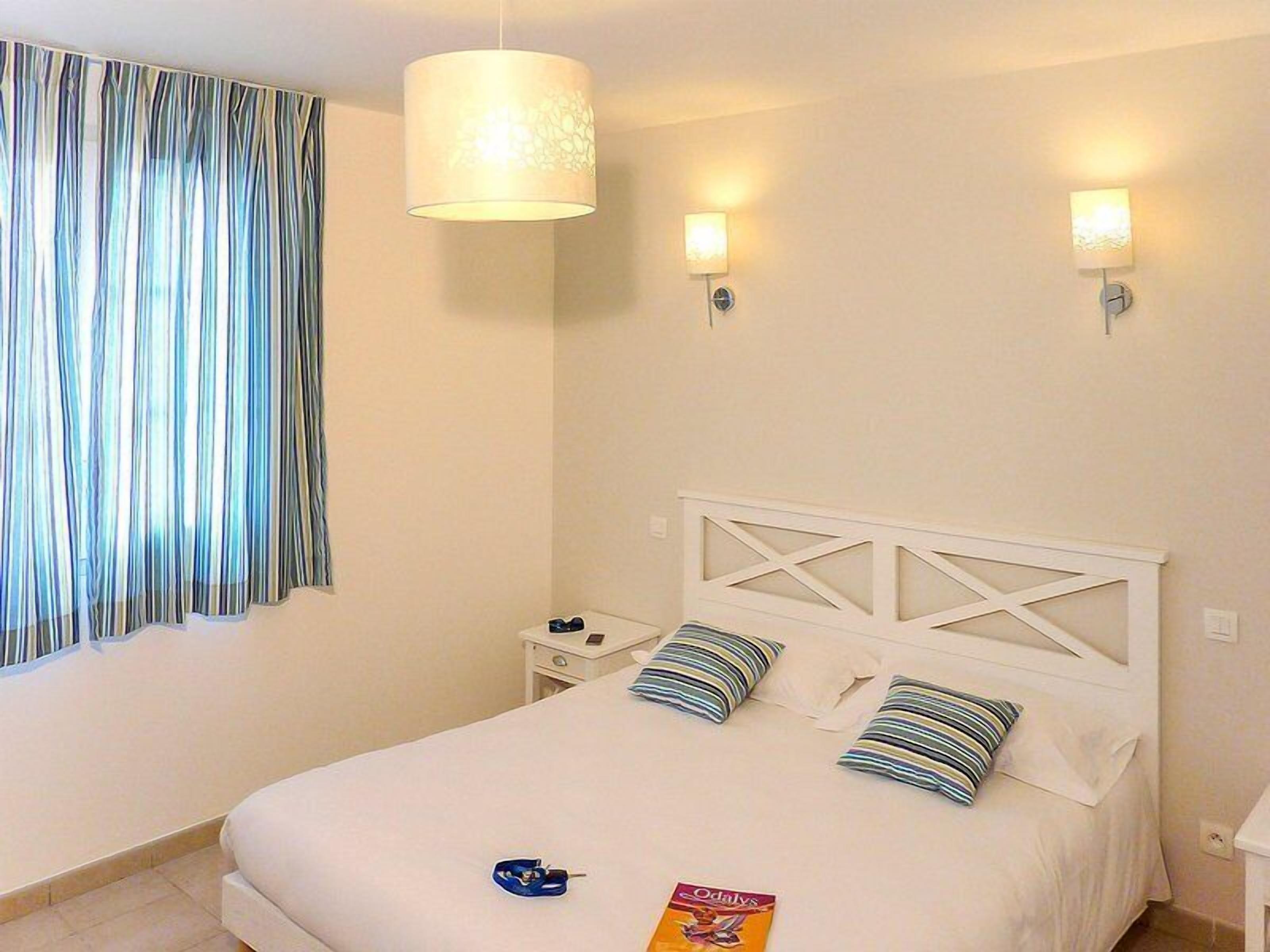 Una habitación de hotel con una cama, dos lámparas de noche, una mesita de noche y una cortina.