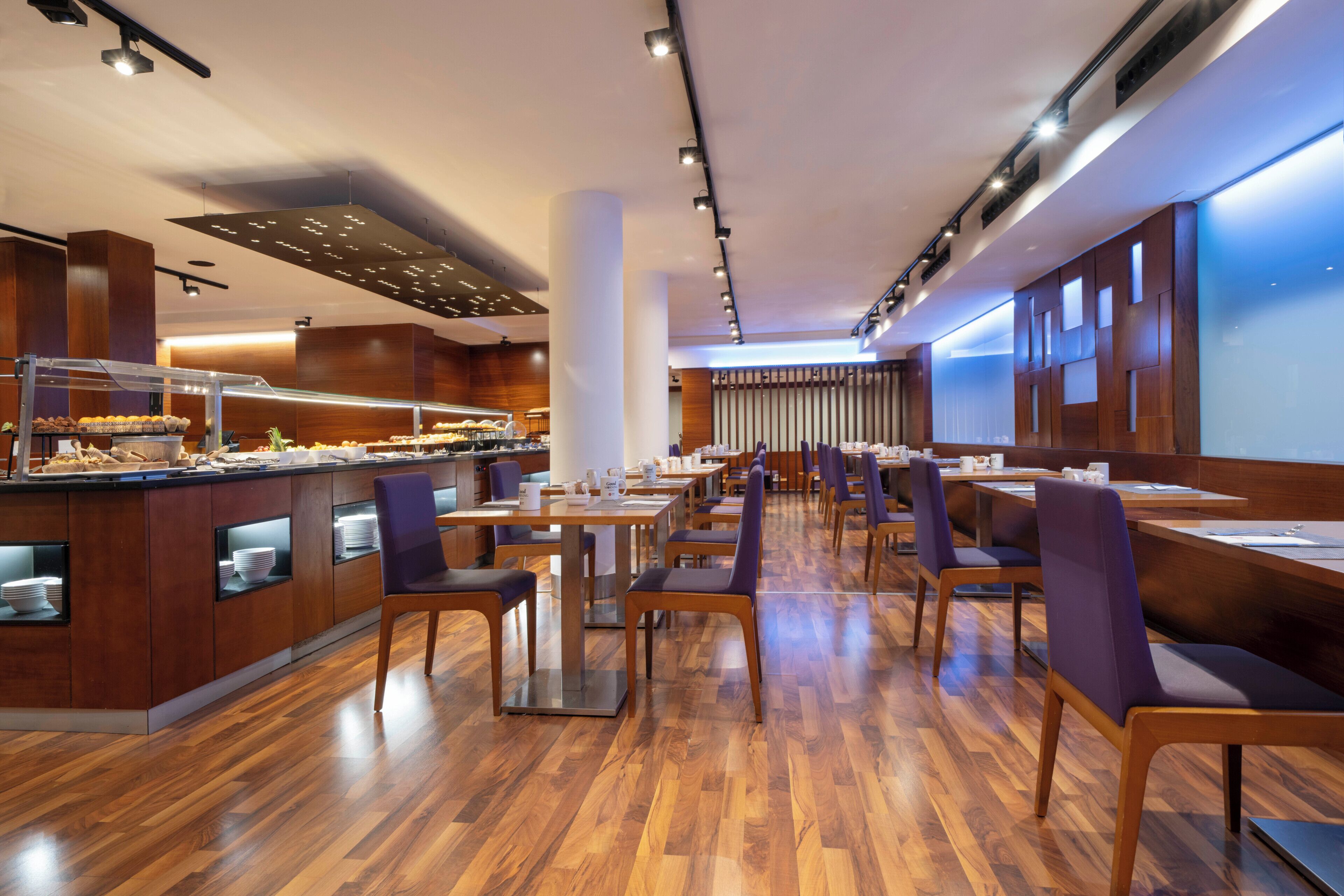 Um restaurante moderno com piso de madeira, área de buffet e sala de jantar com mesas e cadeiras.