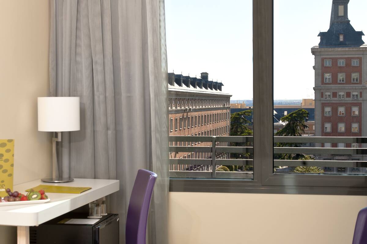 Habitación de hotel con un ventanal grande, un escritorio con silla, una cama y vista a un paisaje urbano con un edificio destacado.