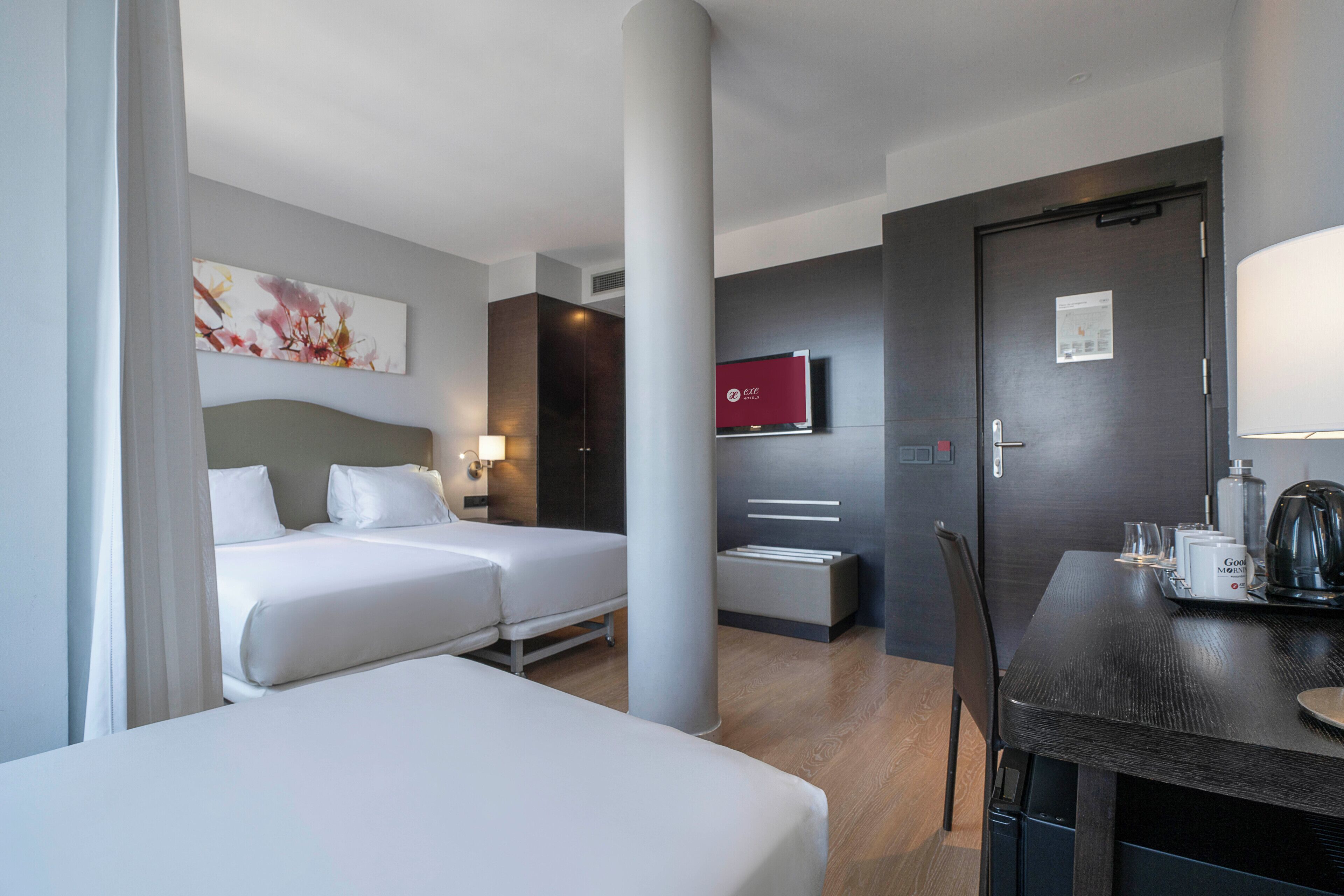 Una habitación de hotel con dos camas, un escritorio con cafetera y un televisor en la pared.