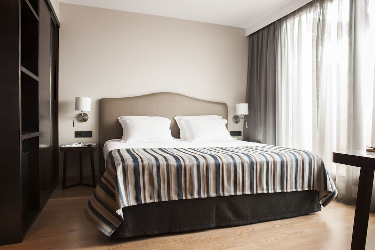 Habitación de hotel con una cama grande, dos lámparas de noche, un armario de madera oscura y un ventanal con cortinas translúcidas.
