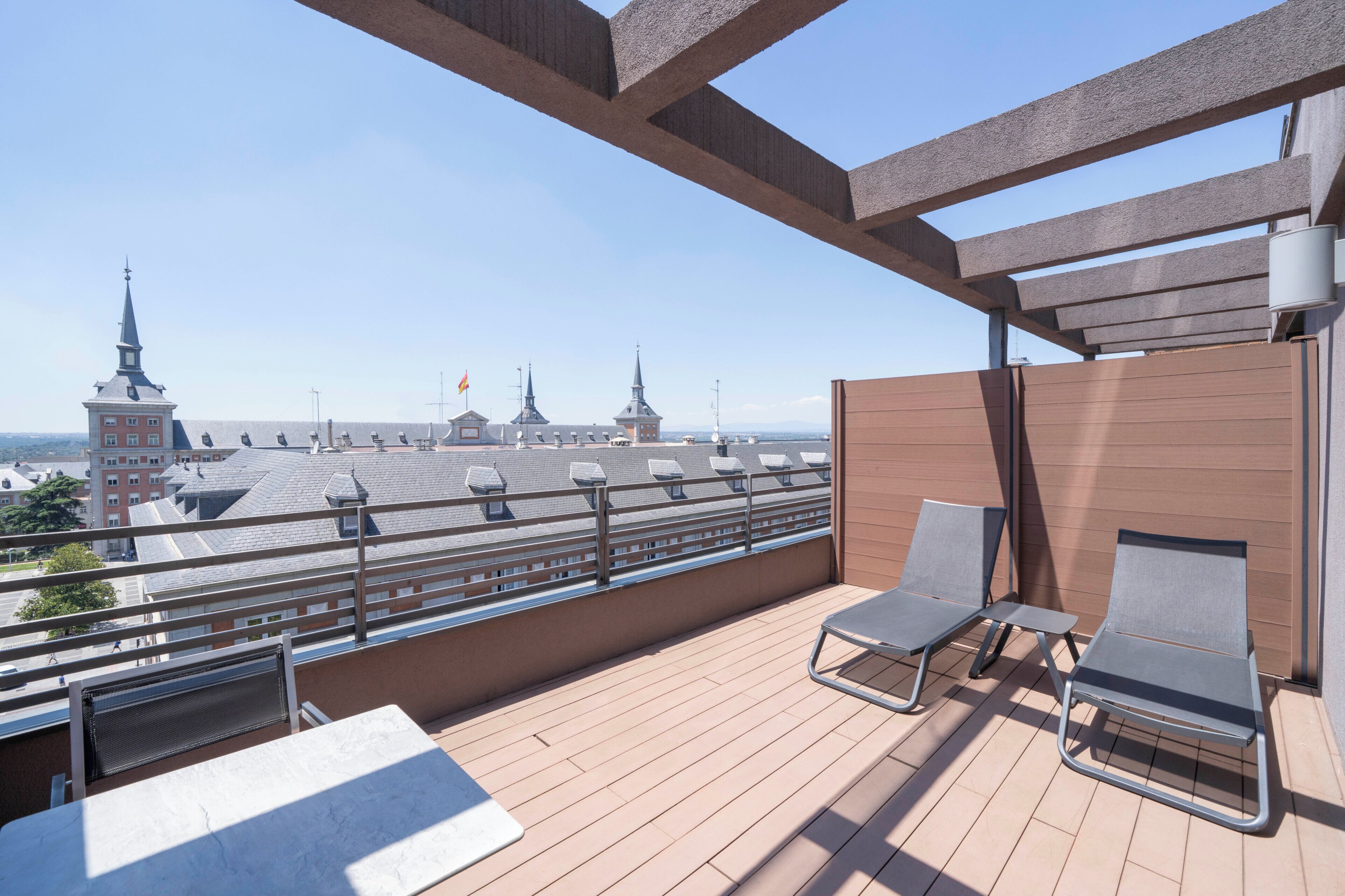 Un balcón con dos sillones de descanso y una mesa, con vistas a un paisaje urbano con varios edificios y agujas.