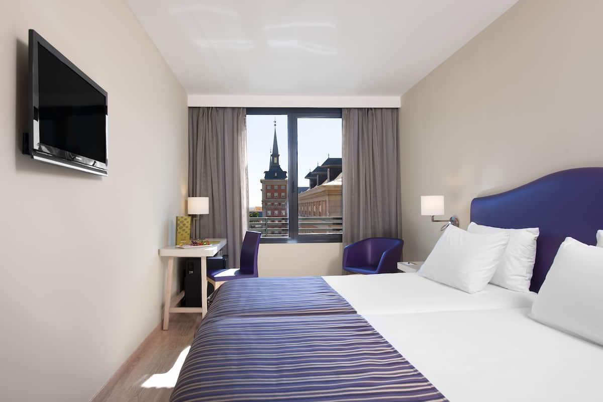 Una habitación de hotel con una cama, un escritorio con una silla, un televisor montado en la pared y una ventana con vista a un edificio.