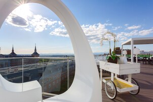 Eine Dachterrasse mit freiem Blick auf Stadtgebäude, ein weißer Wagen mit einer Pflanze und ein sonnendurchfluteter Himmel.