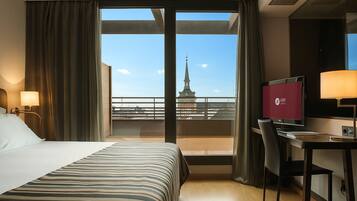 Een hotelkamer met een bed, bureau en uitzicht op een stadsbeeld door grote ramen.