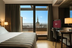 Camera d'albergo con un letto, una scrivania e vista sulla città attraverso ampie finestre.