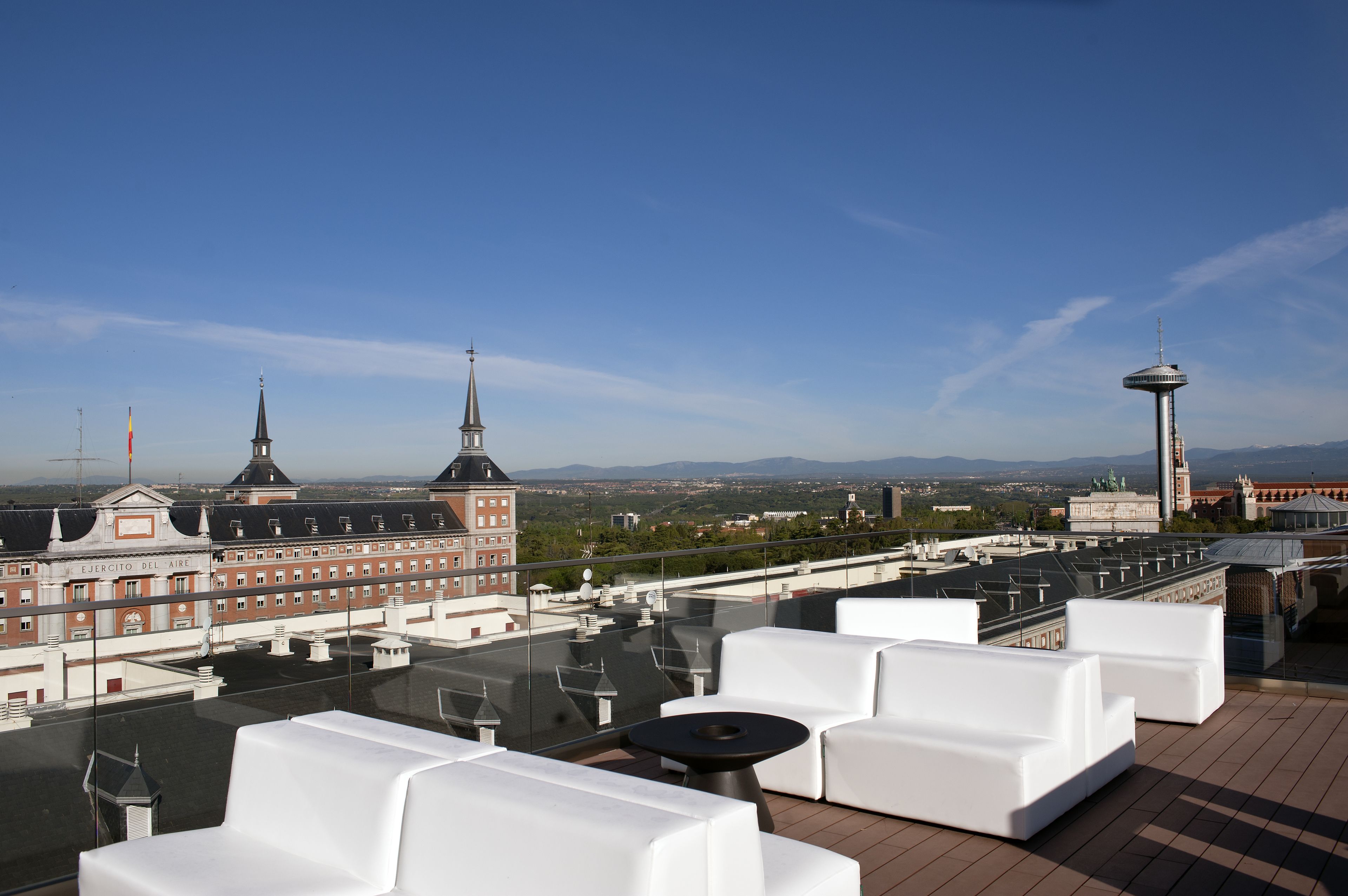 Terraço no telhado com sofás brancos e uma mesa, com vista para o horizonte urbano com edifícios históricos e uma torre.
