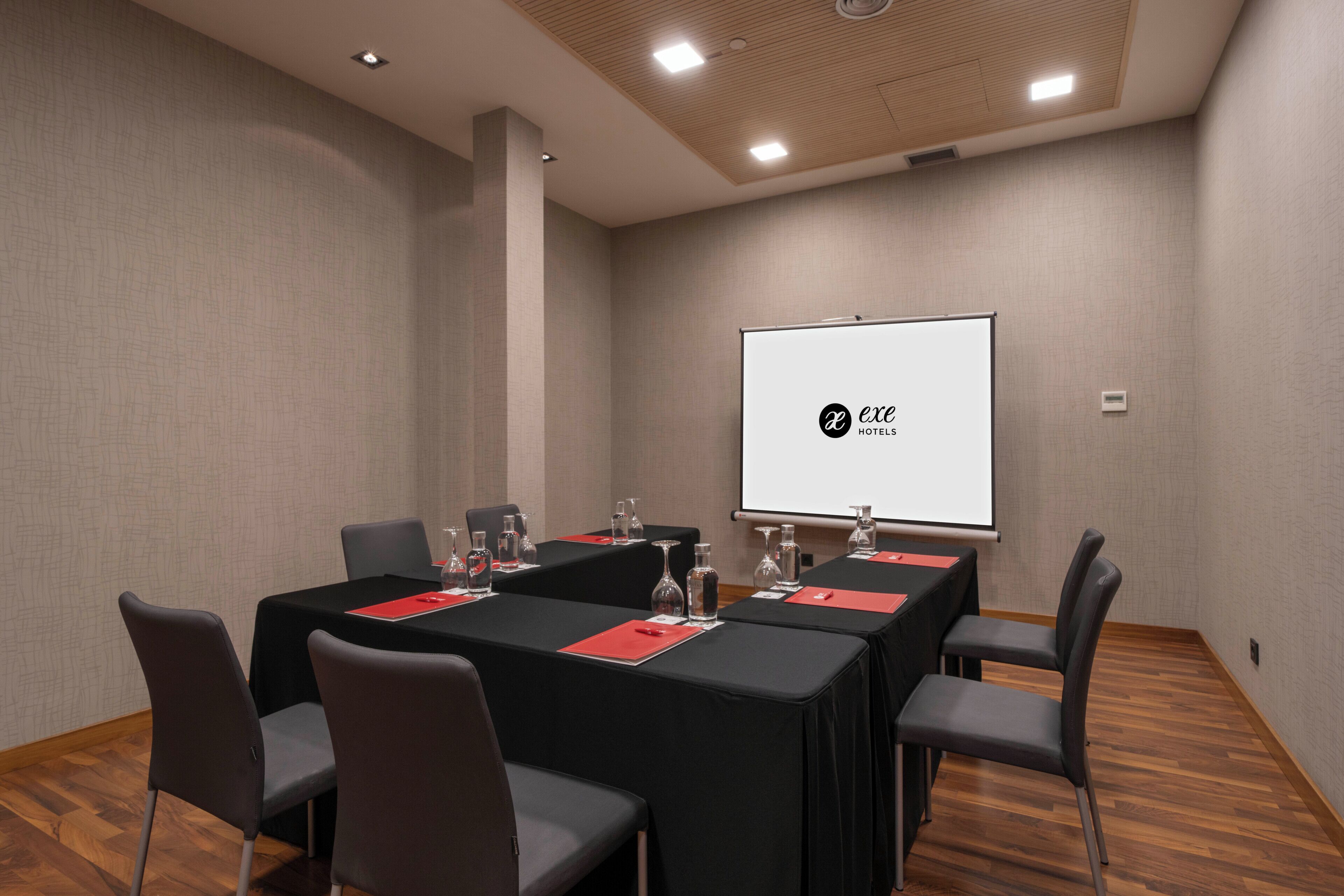 Un salón de conferencias con una larga mesa, sillas, una pantalla de proyector y el logotipo de 'exel hotels'.