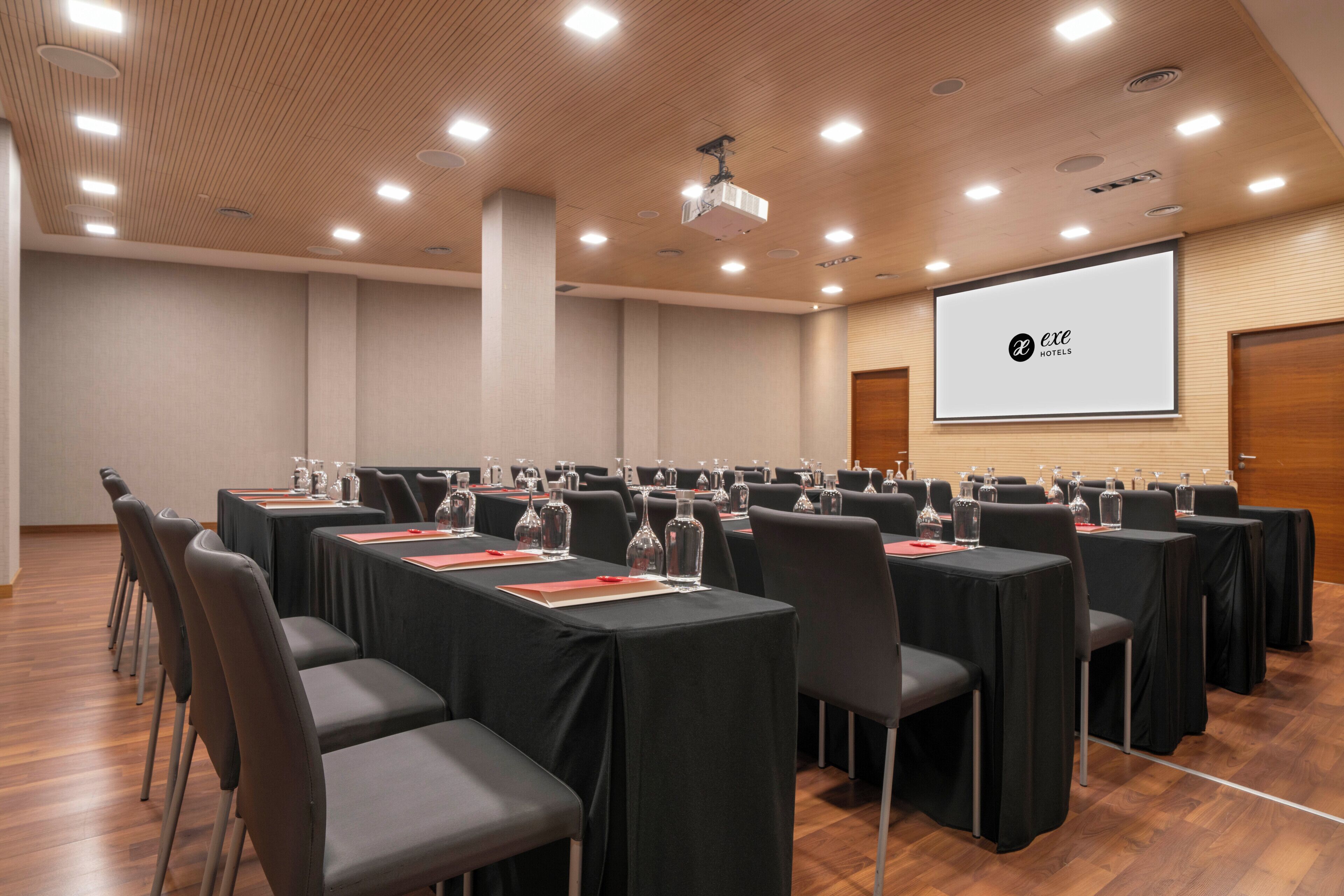 Une salle de conférence avec des rangées de tables, des chaises et un écran de projection affichant un logo.