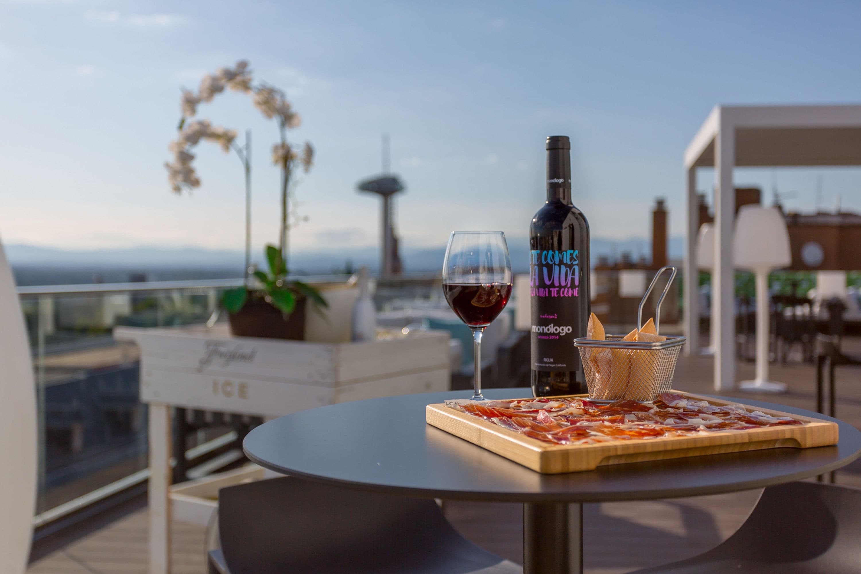 Een tafel met pizza, een fles wijn en een glas op een terras op het dak, met uitzicht op een toren.