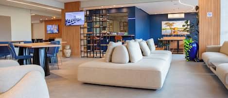 A modern hotel lobby with a seating area, a television, a reception desk, and a bookshelf.