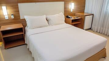 Habitación de hotel con una cama grande, cabecera de madera, mesitas de noche, un pequeño refrigerador y una ventana con cortinas.