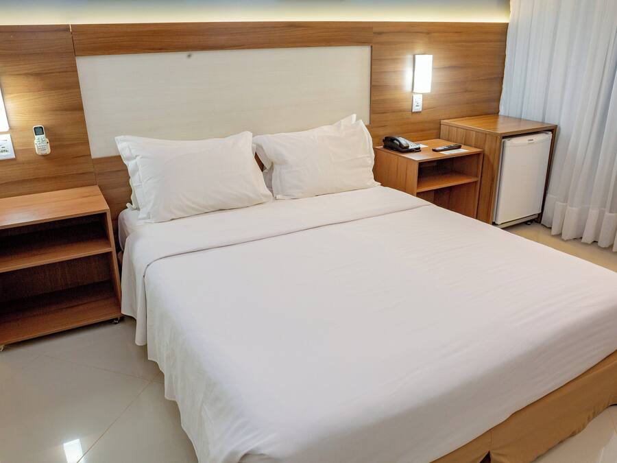 Habitación de hotel con una cama grande, cabecera de madera, mesitas de noche, un pequeño refrigerador y una ventana con cortinas.