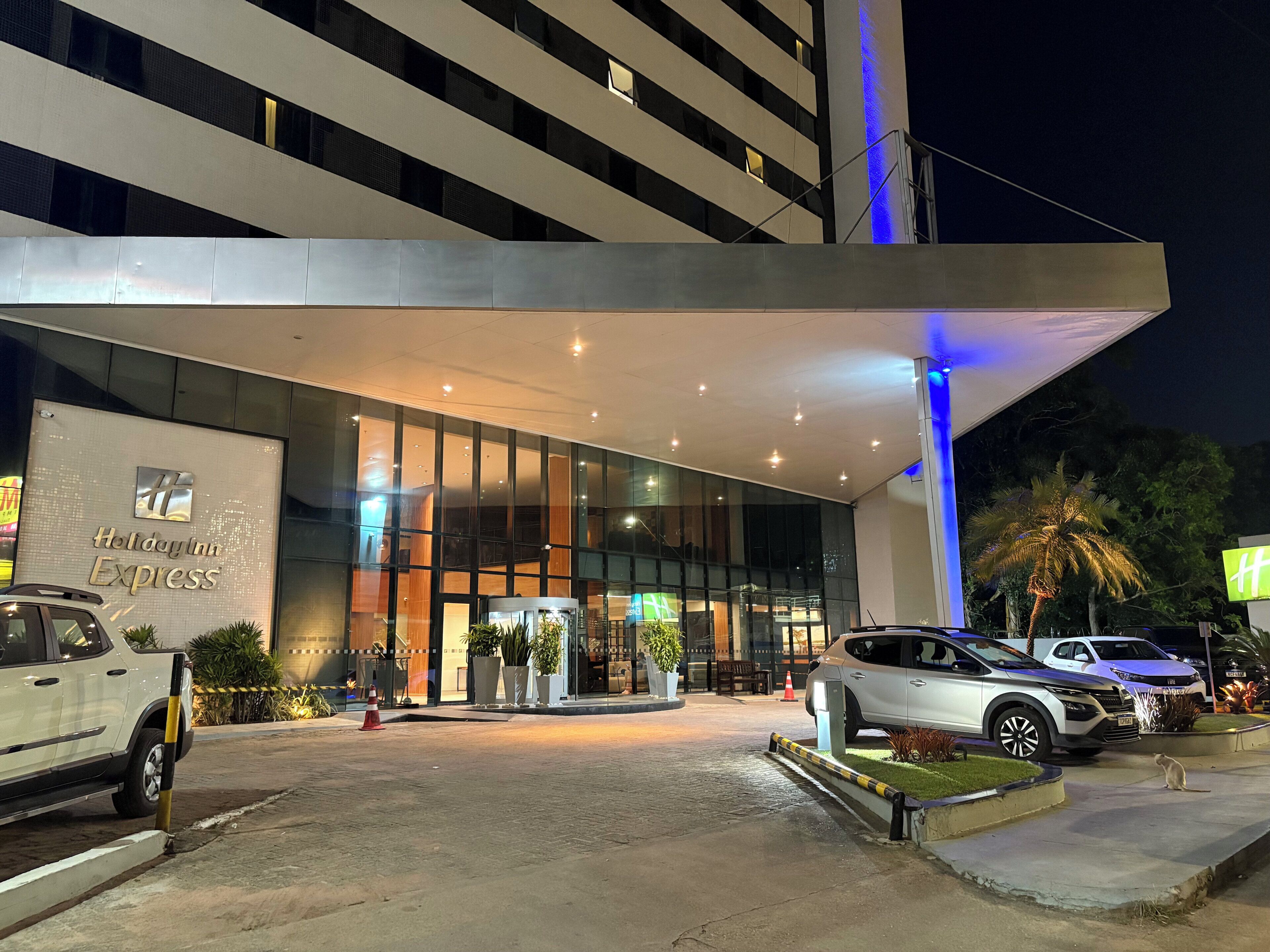 Entrada de un hotel moderno con fachada de vidrio, una galería techada y un estacionamiento con automóviles.