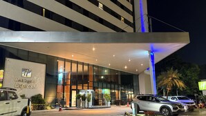 Une entrée d’hôtel moderne avec une façade en verre, une allée couverte et un espace de stationnement avec des voitures.