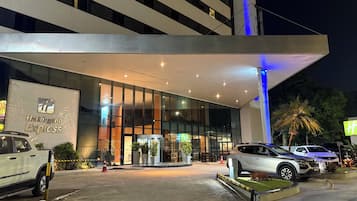 Entrada de un hotel moderno con fachada de vidrio, una galería techada y un estacionamiento con automóviles.