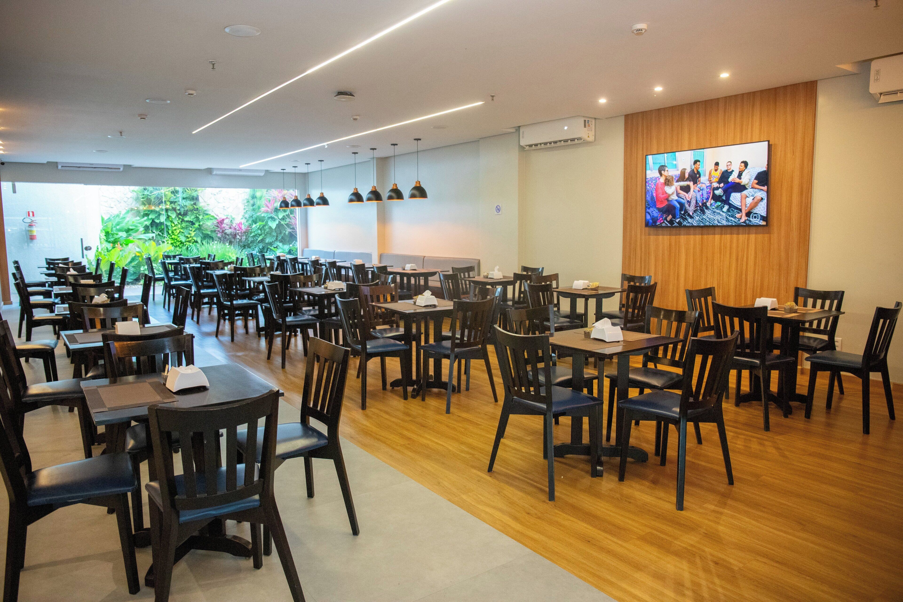 A spacious restaurant with multiple tables and chairs, a large screen on the wall, and a view of greenery outside.