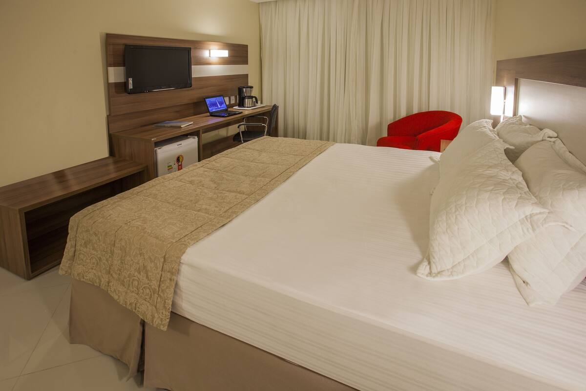 Une chambre d’hôtel avec un grand lit, un bureau avec un ordinateur portable, une chaise rouge et un téléviseur fixé au mur.