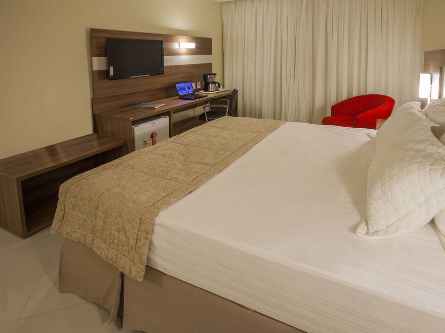A hotel room with a large bed, a desk with a laptop, a red chair, and a television on the wall.