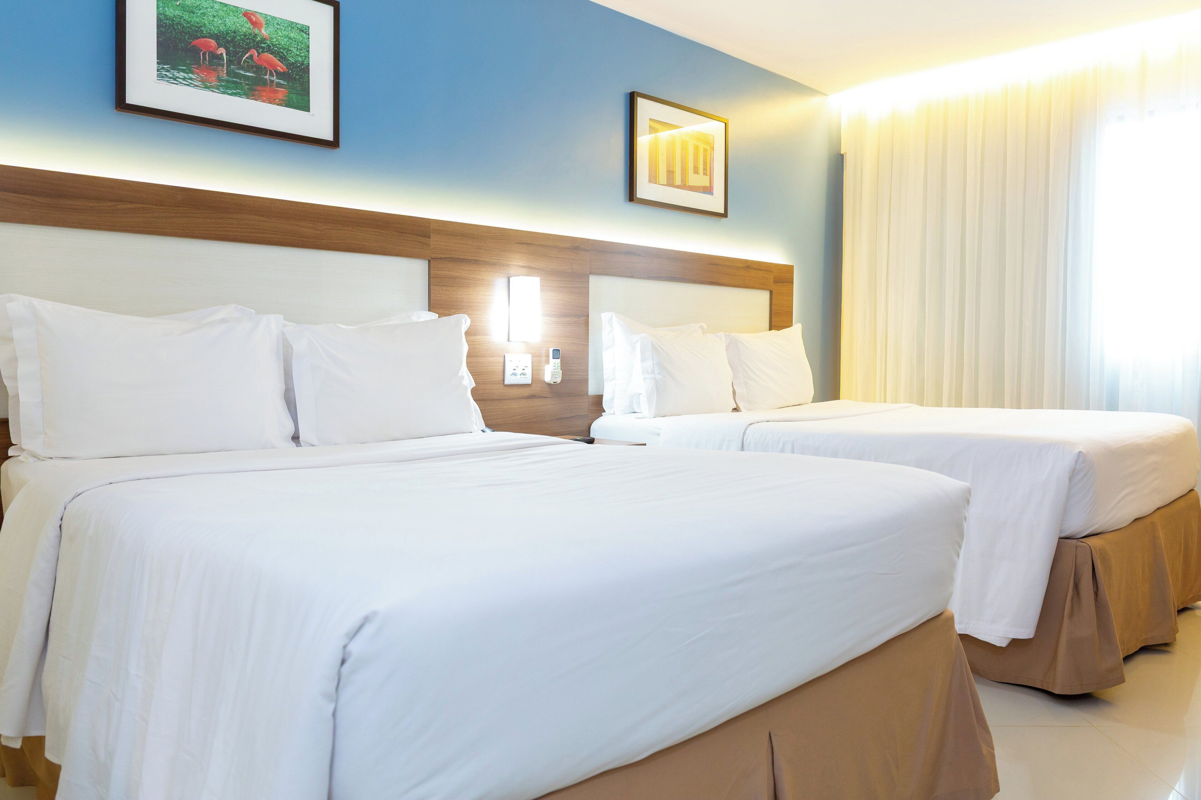 Habitación de hotel con dos camas, cabecero, mesita de noche con lámpara y ventana con cortinas.