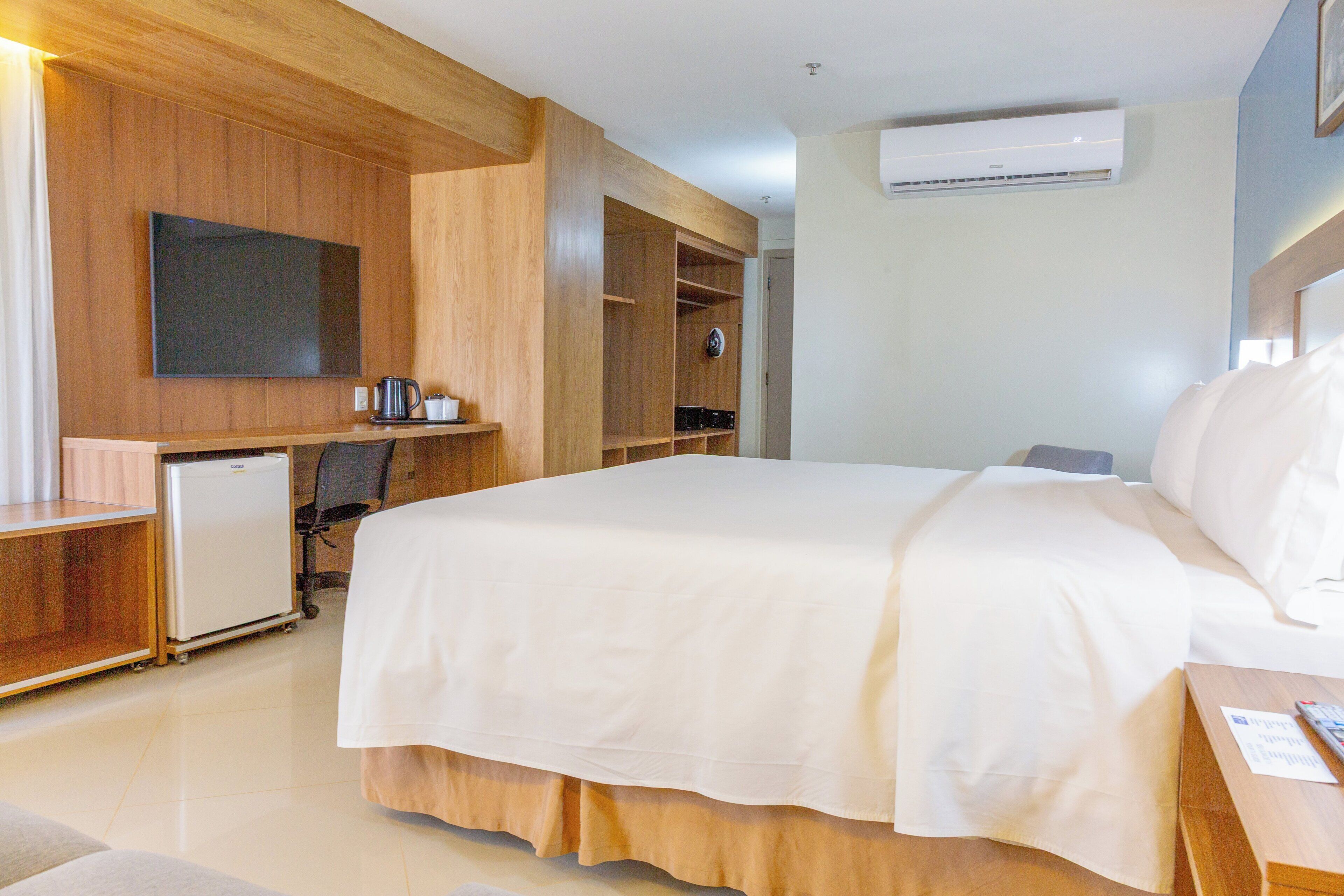 A hotel room with a large bed, a desk, a television, a mini-fridge, and a wardrobe.