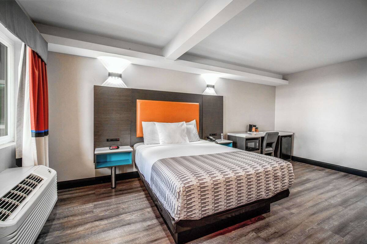 A modern hotel room with a large bed, a desk, and a chair. There is a heater on the floor and a window with curtains.