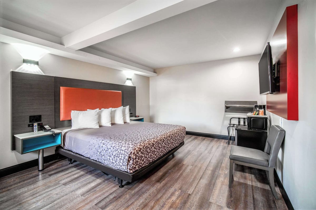 A modern hotel room with a large bed, a desk with a chair, a flat-screen TV mounted on the wall, and a small kitchenette area.